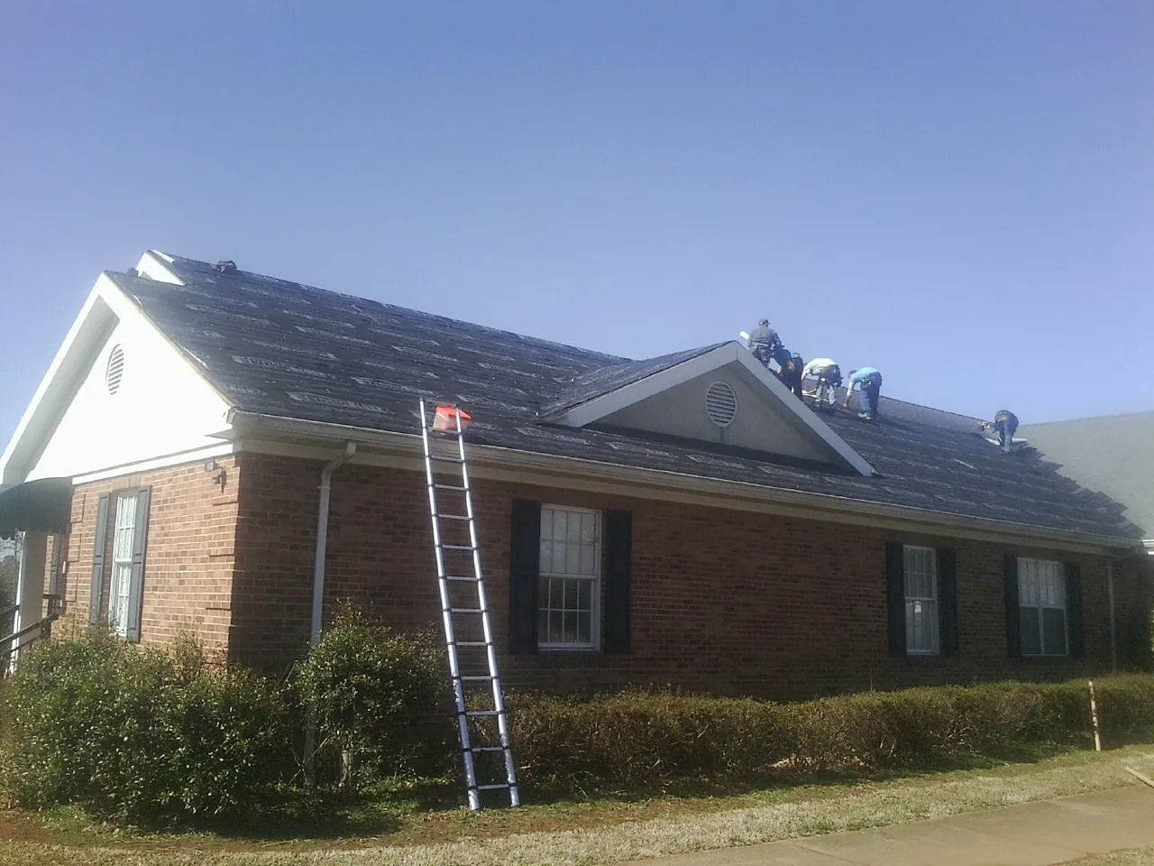 Crew tearing shingles off a brick ranch roof with a ladder set — Metal Roof Repair work by our crew in Hensley