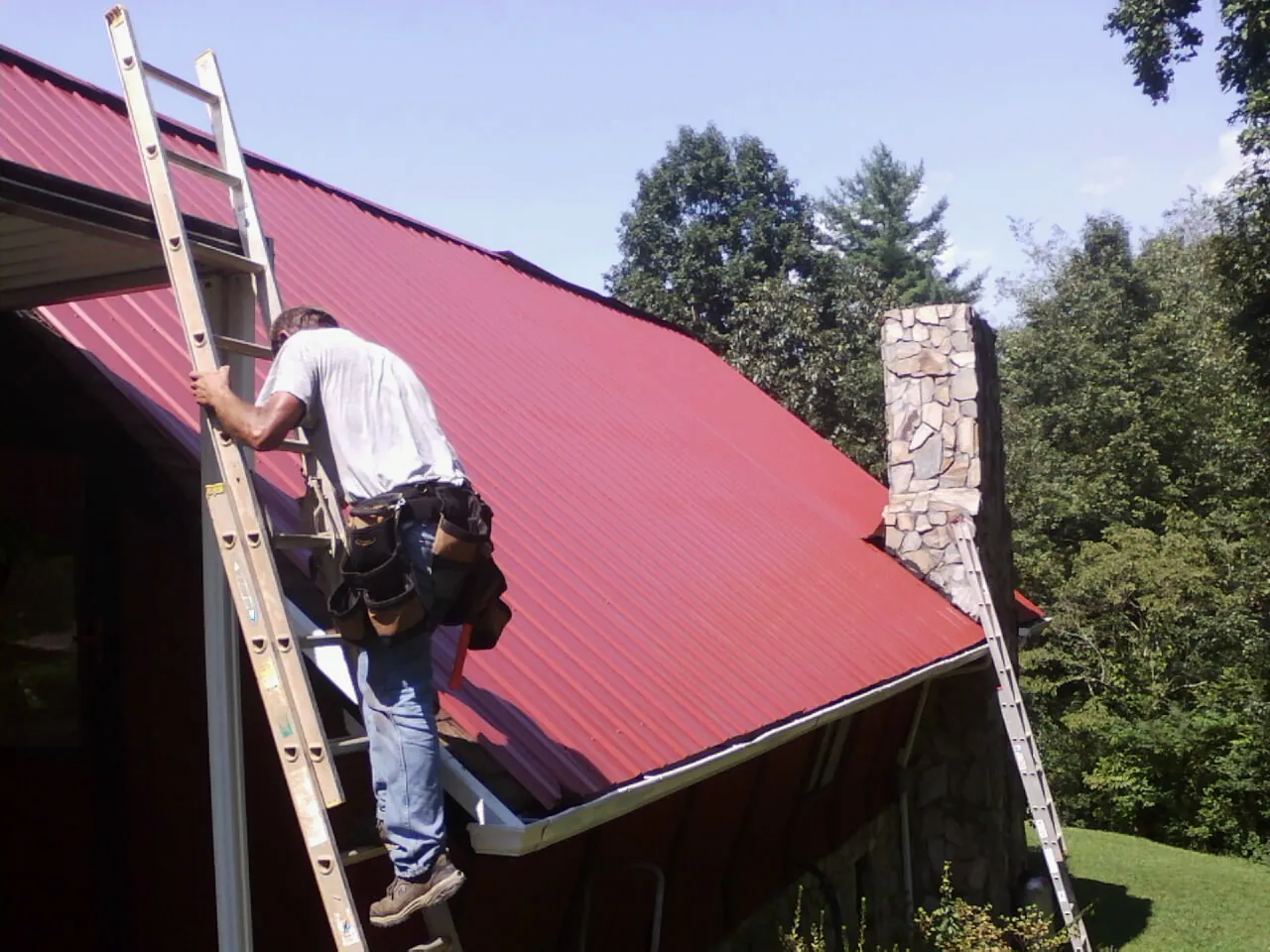 Roofer on a ladder beside a red metal roof and stone chimney — Soffit & Fascia Repair by Roofing Brothers in Hensley
